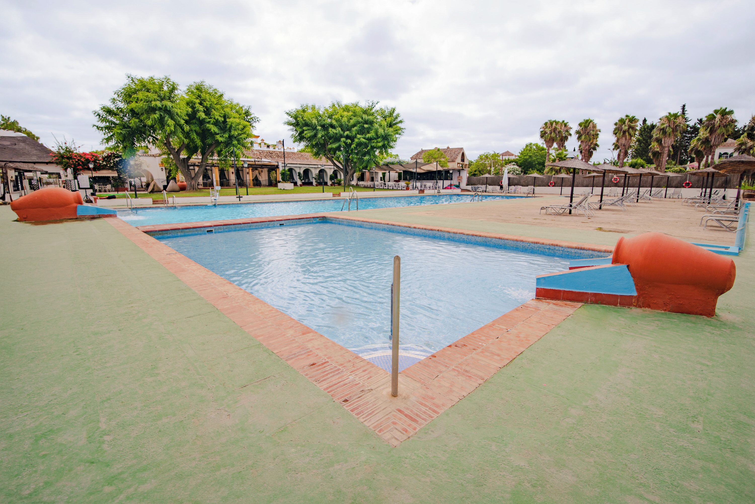 Una piscina con borde de baldosas, rodeada de palmeras y una zona cubierta con sillones de descanso.