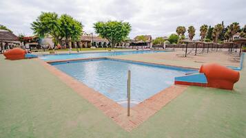 Una piscina con borde de baldosas, rodeada de palmeras y una zona cubierta con sillones de descanso.