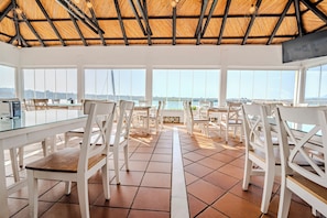A spacious dining area with white chairs and tables, large windows offering a view of the sea, and a thatched roof ceiling. - La Molinera Bungalows (Arcos de la Frontera)