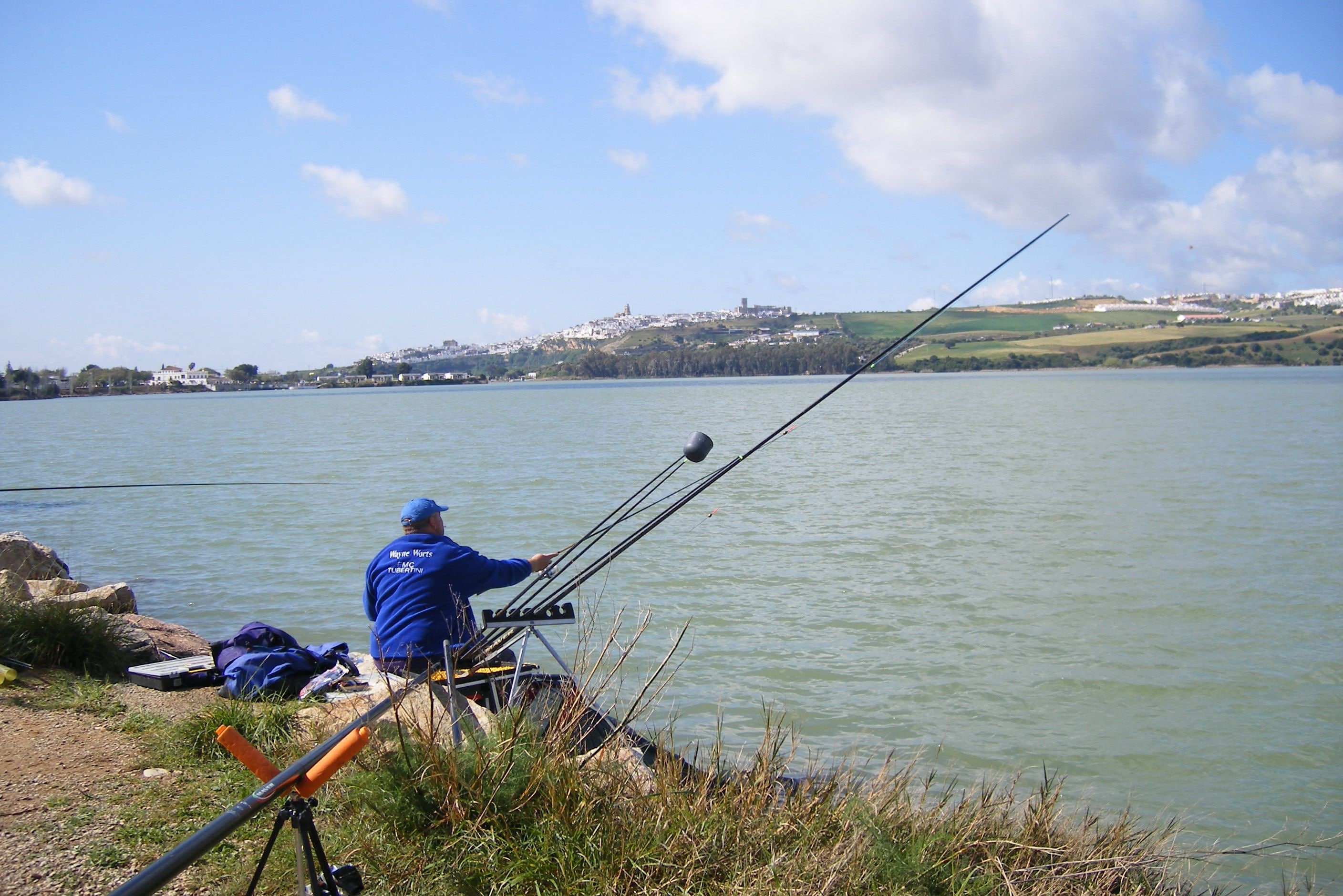 Una persona pescando en un lago tranquilo, con vista despejada a un pueblo distante en la orilla opuesta.