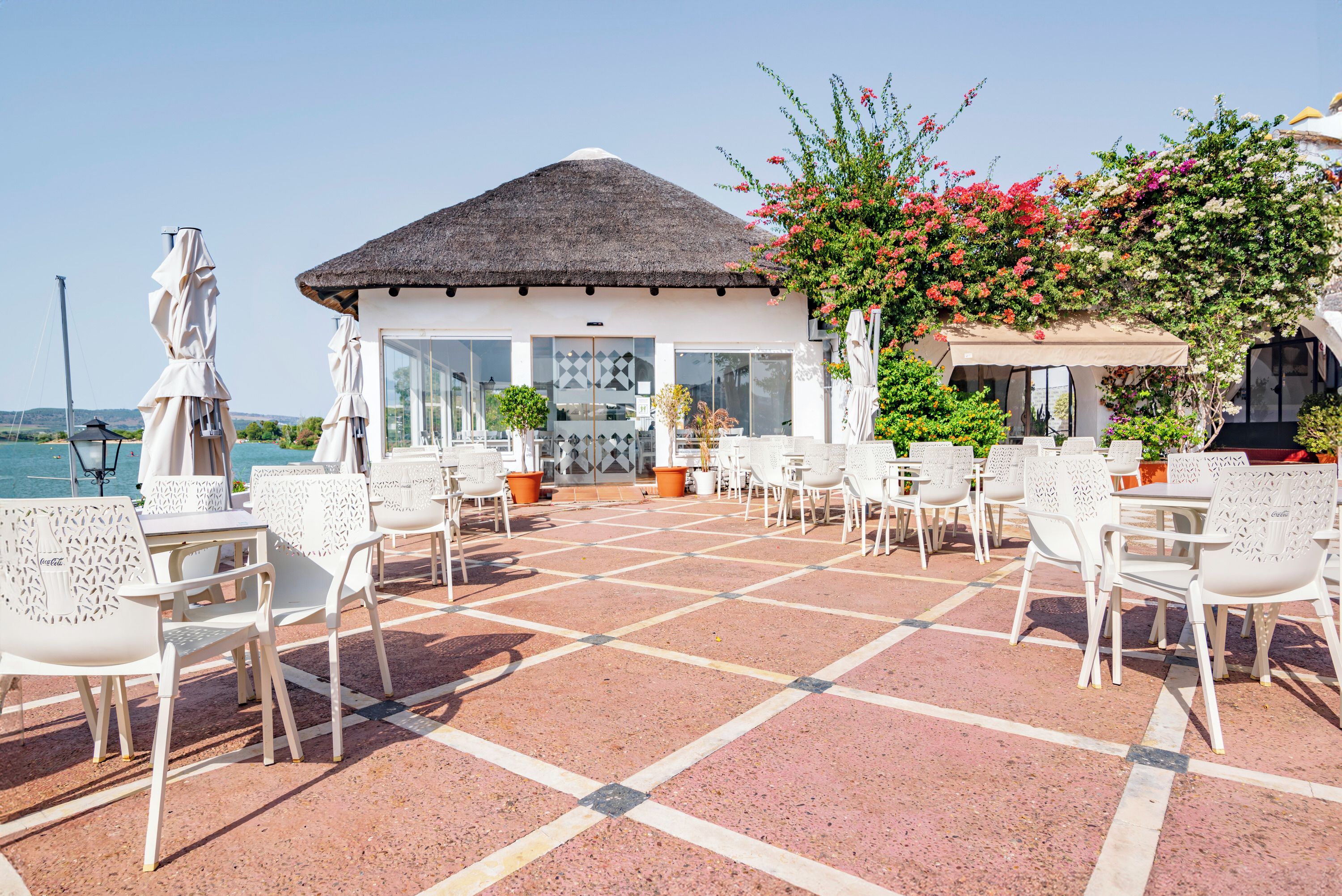 A terrace with white chairs and tables, a thatched-roof building, and a view of the sea.