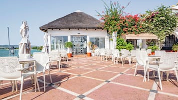 A terrace with white chairs and tables, a thatched-roof building, and a view of the sea.