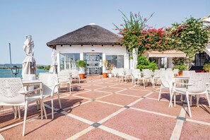Una terraza con sillas y mesas blancas, un edificio con techo de paja y vista al mar.