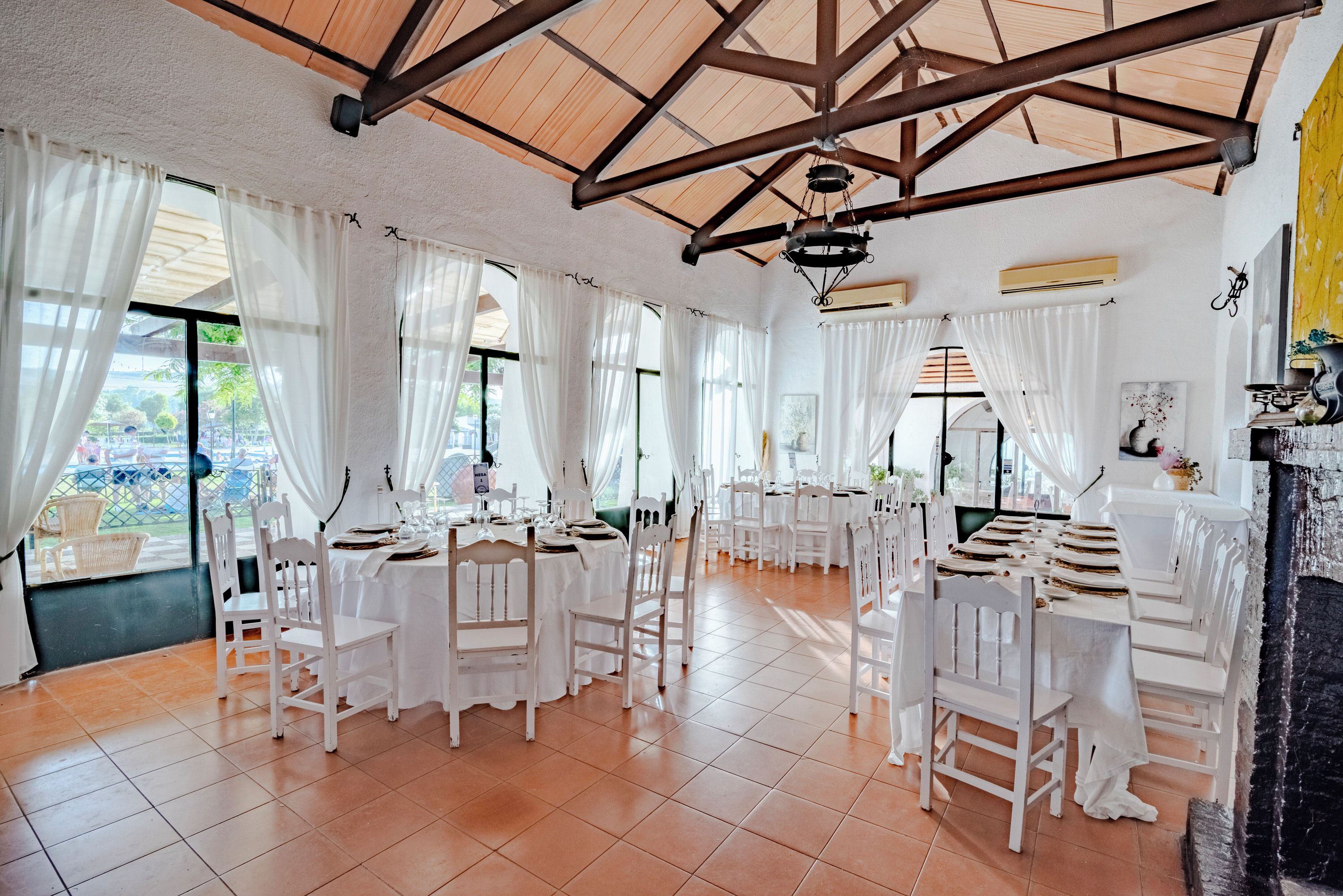 Un comedor amplio con mesas y sillas blancas, ventanas grandes con cortinas blancas y un techo alto de madera.