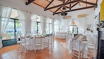 Un comedor amplio con mesas y sillas blancas, ventanas grandes con cortinas blancas y un techo alto de madera.