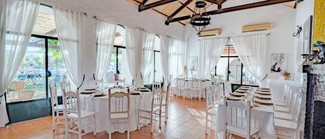 Un vaste espace dédié à la salle à manger, avec des tables et des chaises blanches, de grandes fenêtres ornées de rideaux blancs et un haut plafond en bois.
