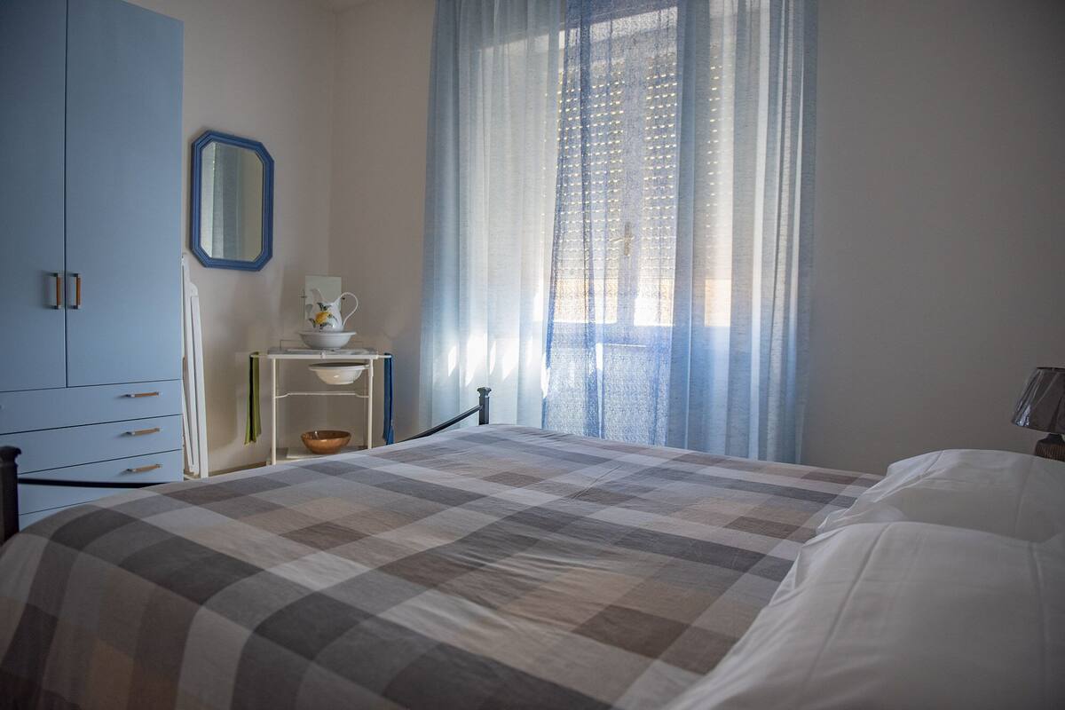 A bedroom with a bed, a dresser, a nightstand, a mirror, and a window with sheer curtains.