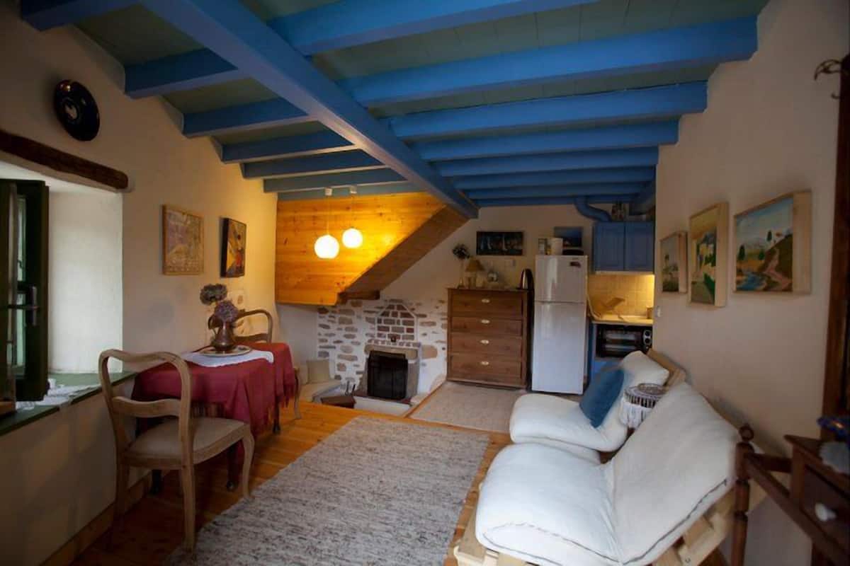 A cozy living room with a fireplace, wooden ceiling beams, a dining area with a table and chairs, and a kitchen area with cabinets and a refrigerator.