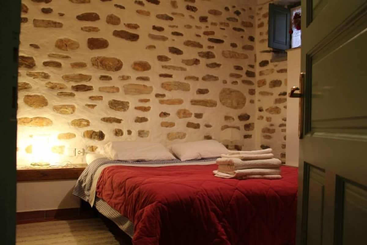 A bedroom with a stone wall, a bed with a red blanket, and a bedside lamp.