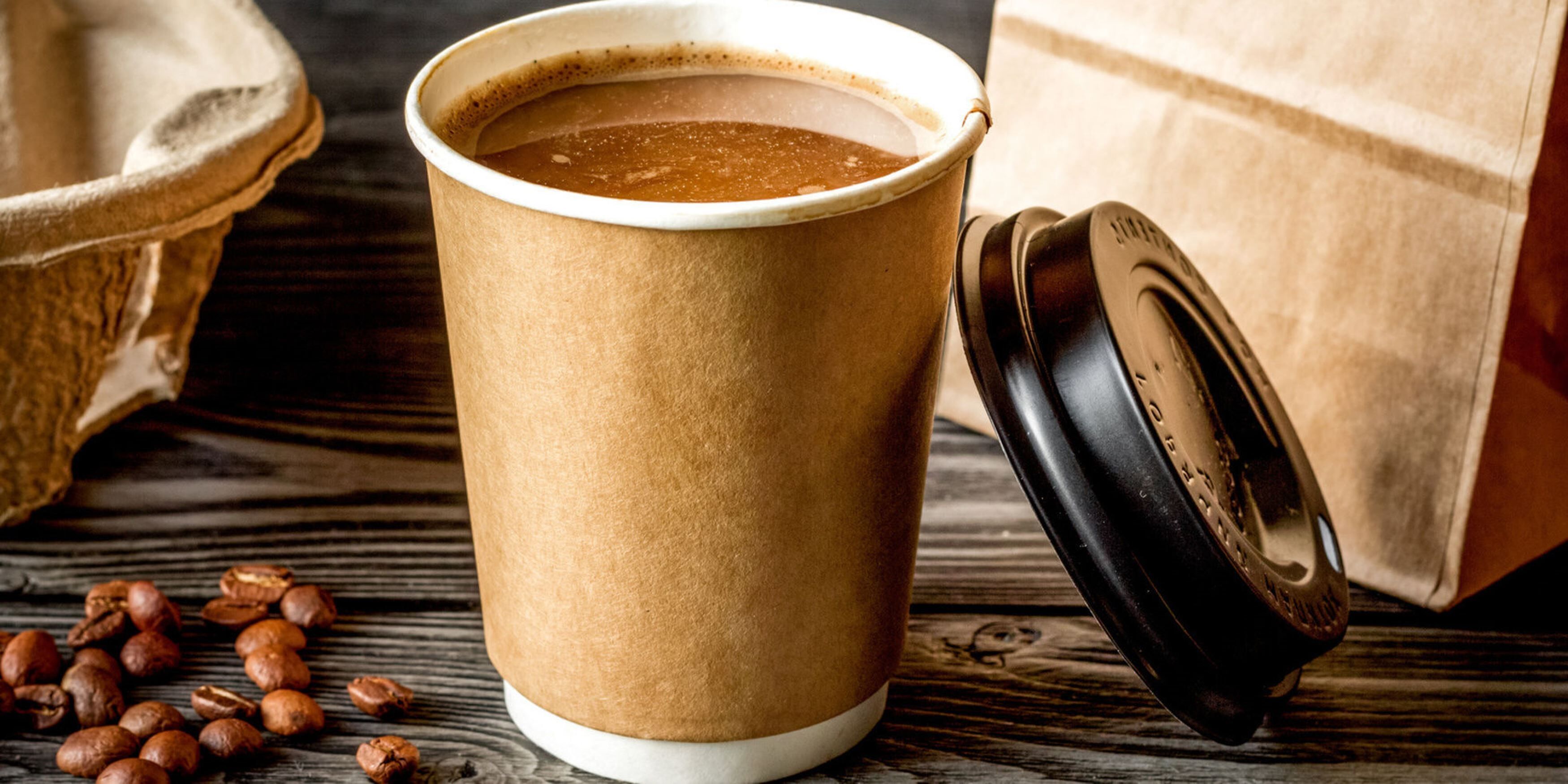 A paper coffee cup with a lid on a wooden surface, surrounded by coffee beans.