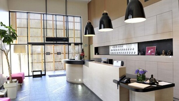 A modern hotel lobby with a reception desk, large windows with blinds, and a seating area with a plant. - Loire & Sens (Les Garennes-sur-Loire)