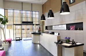 A modern hotel lobby with a reception desk, large windows with blinds, and a seating area with a plant.