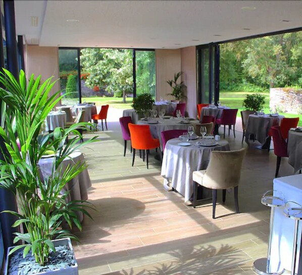 A spacious outdoor dining area with multiple tables and chairs, surrounded by large windows offering a view of a lush garden. - Loire & Sens (Les Garennes-sur-Loire)