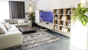 A modern living room with a sectional sofa, a flat-screen TV, a wooden bookshelf, and a large potted plant. - Paramount Bay Penthouse (Birzebbuga)