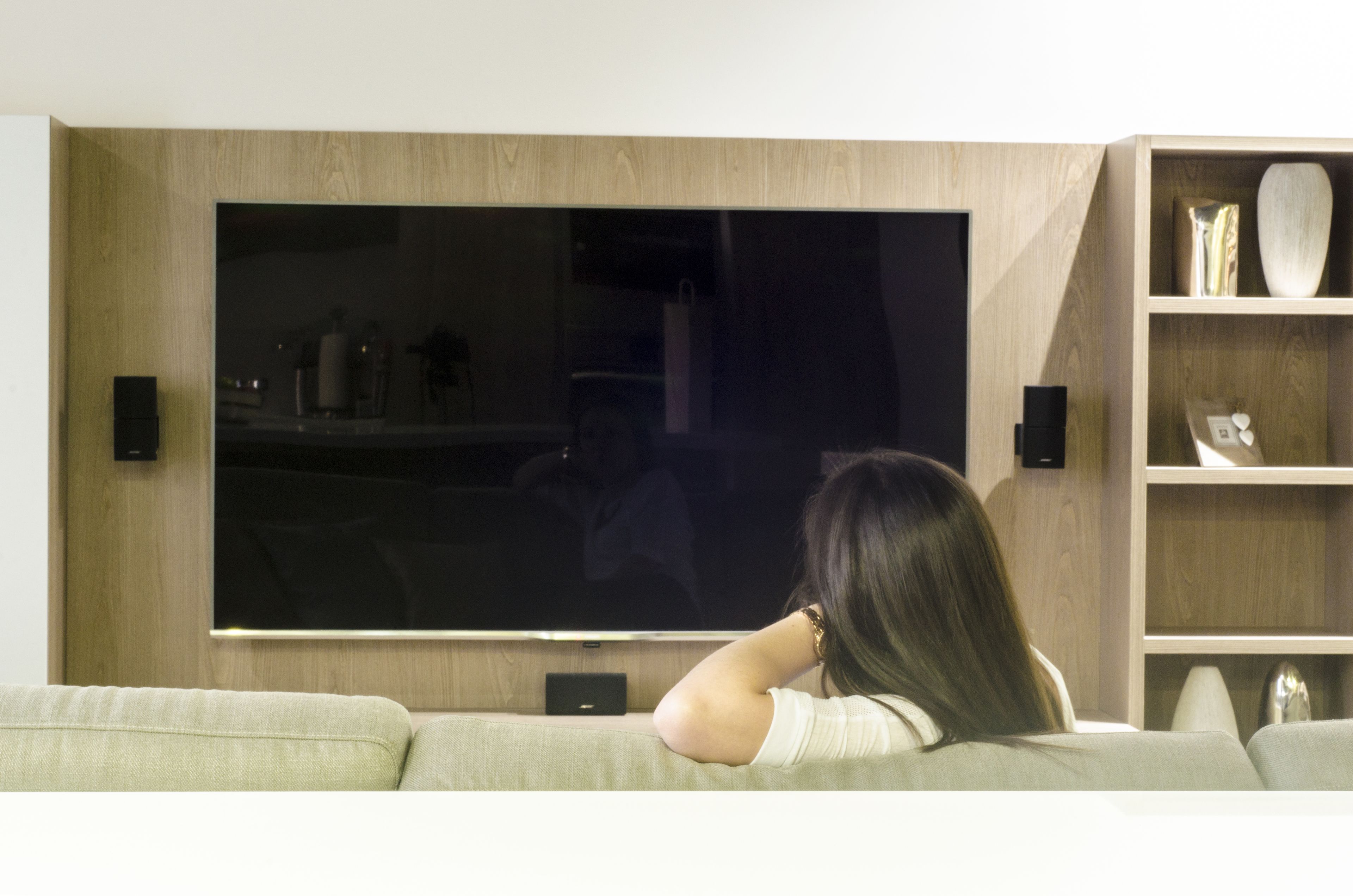 Une femme est assise sur un canapé, regardant la télévision dans un salon moderne, avec des étagères en bois et un téléviseur à écran plat.