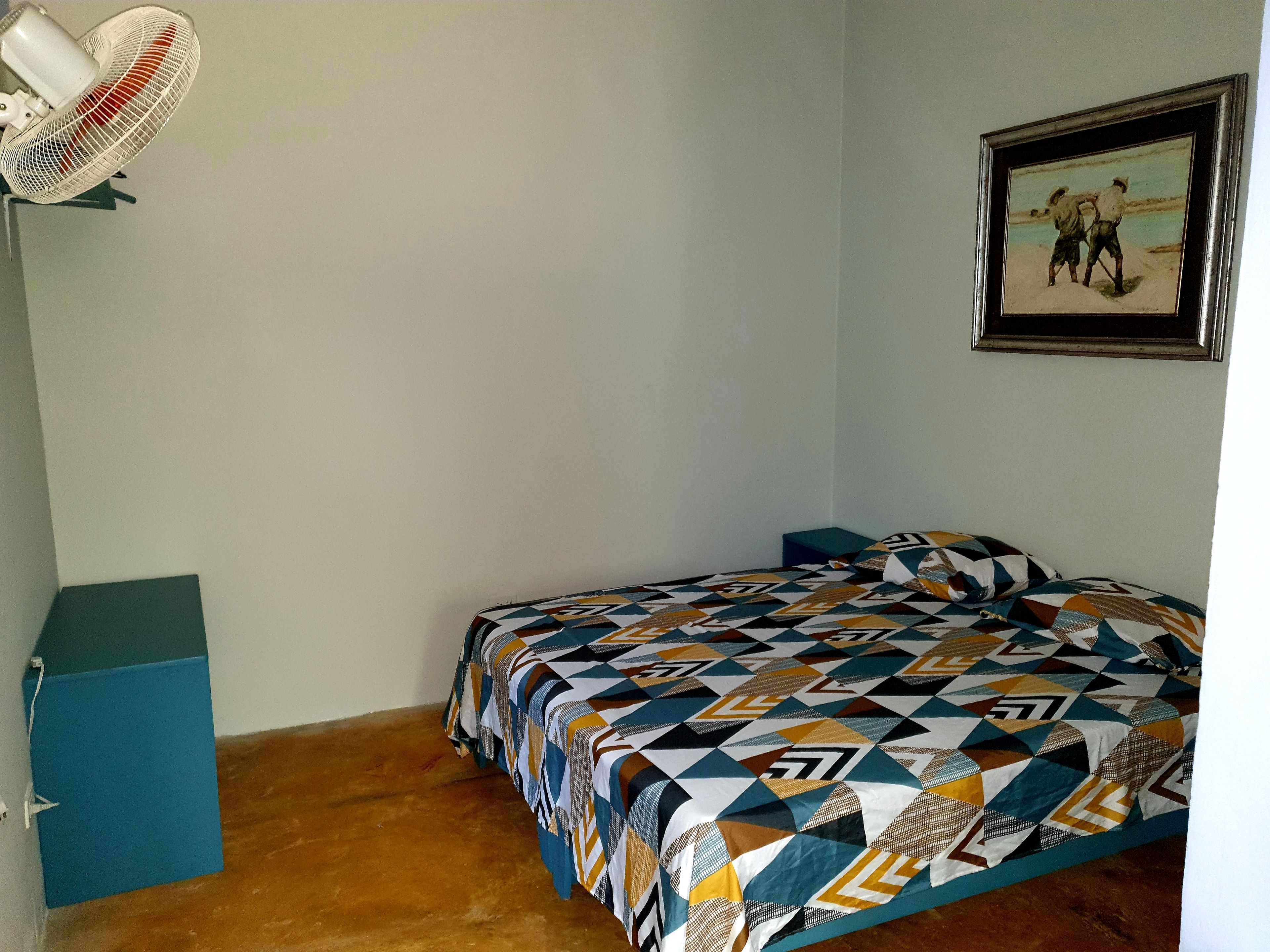 A small, sparsely furnished room with a bed, a standing fan, a framed picture on the wall, and a blue cabinet.