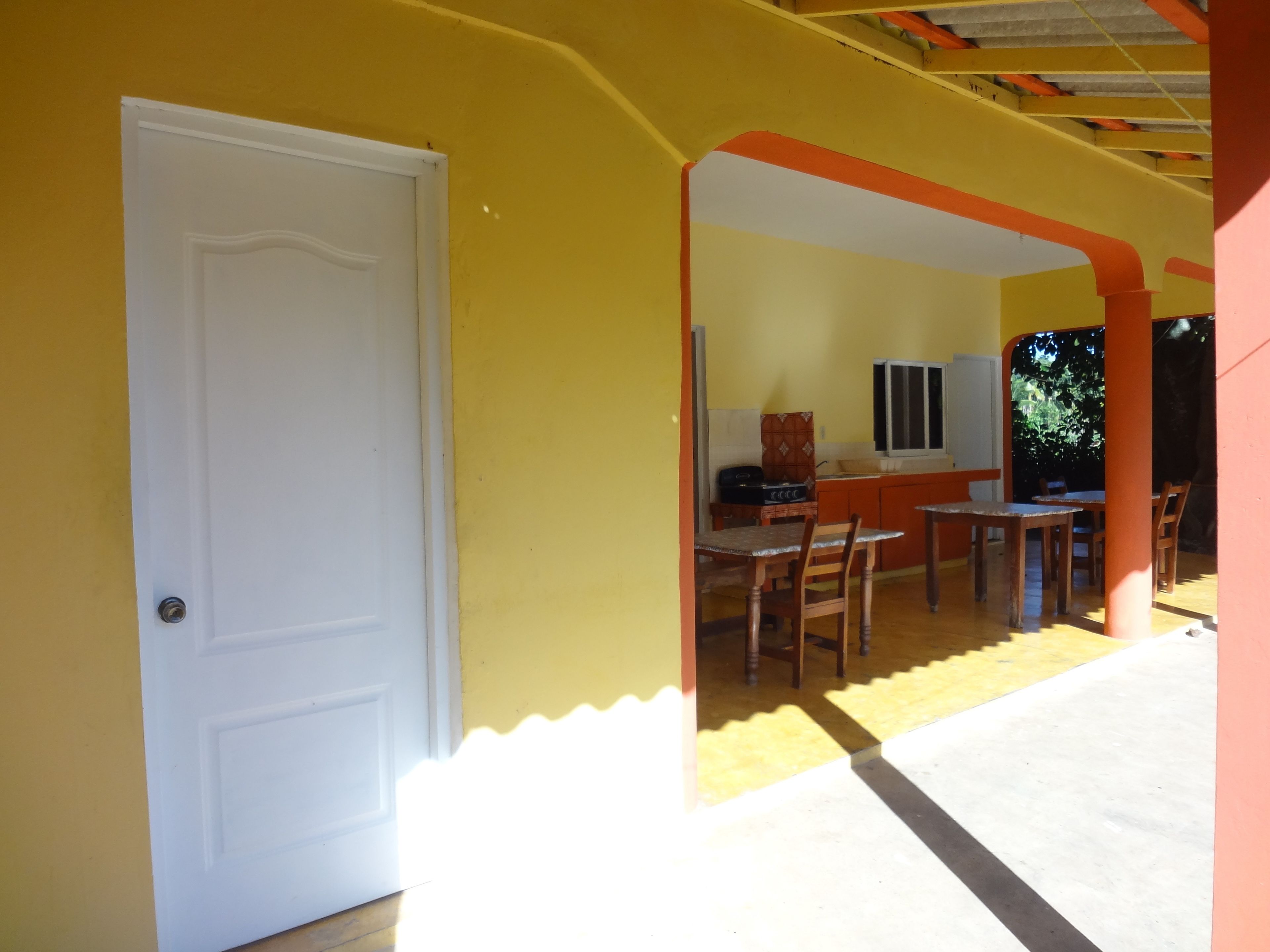 A room with a white door, a dining area with wooden chairs and a table, and a kitchen area with a window and cabinets.