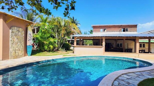 A swimming pool with a stone border, a covered patio area, and a building with a stone wall and a sign indicating 'HOSTEL ROOMS'. - Gipsy Ranch Rooms - Hostel (Cabarete)