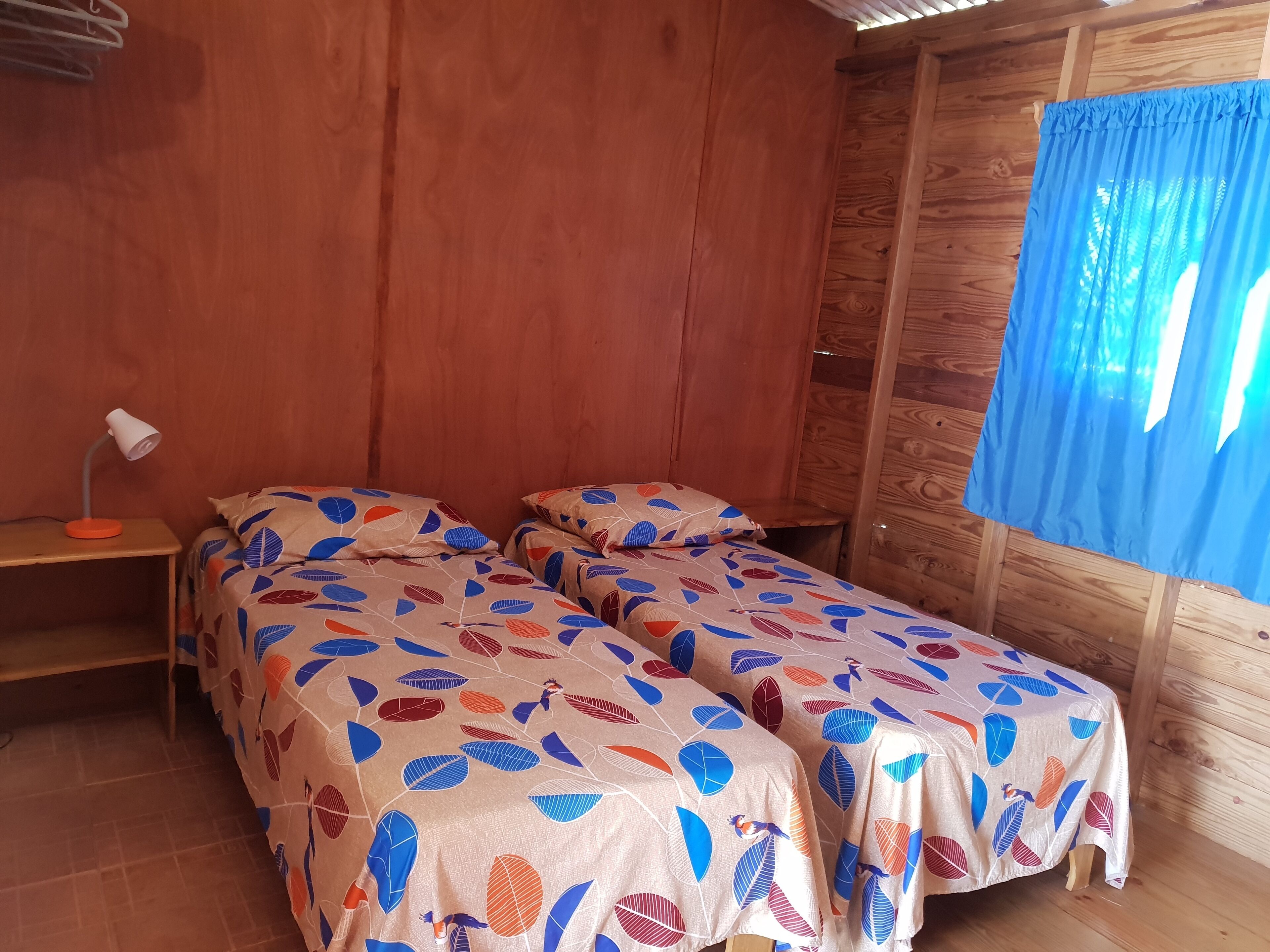 Ein Zimmer mit zwei Betten, Holzwänden, einem kleinen Tisch mit Lampe und einem blauen Vorhang.