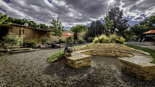 A patio area with stone seating and a pergola, surrounded by greenery and a building with a red roof.