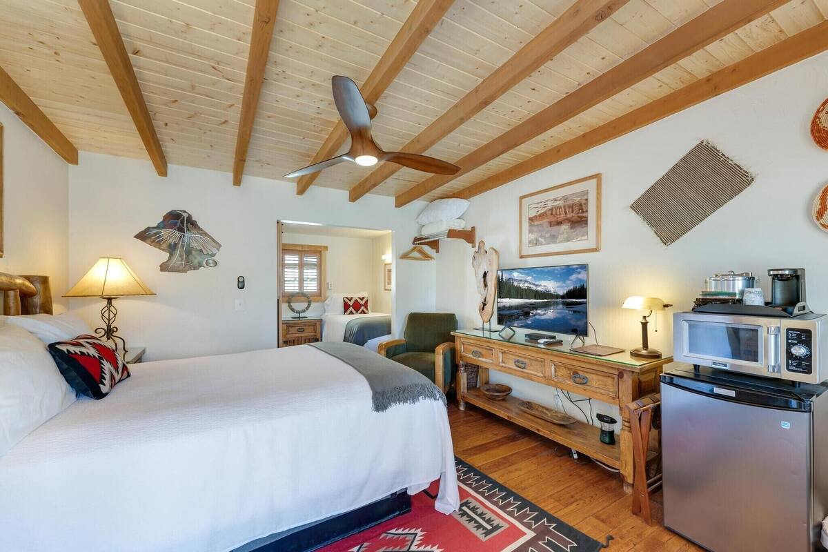 A bedroom with a bed, a desk, a refrigerator, a microwave, a ceiling fan, and a painting on the wall.