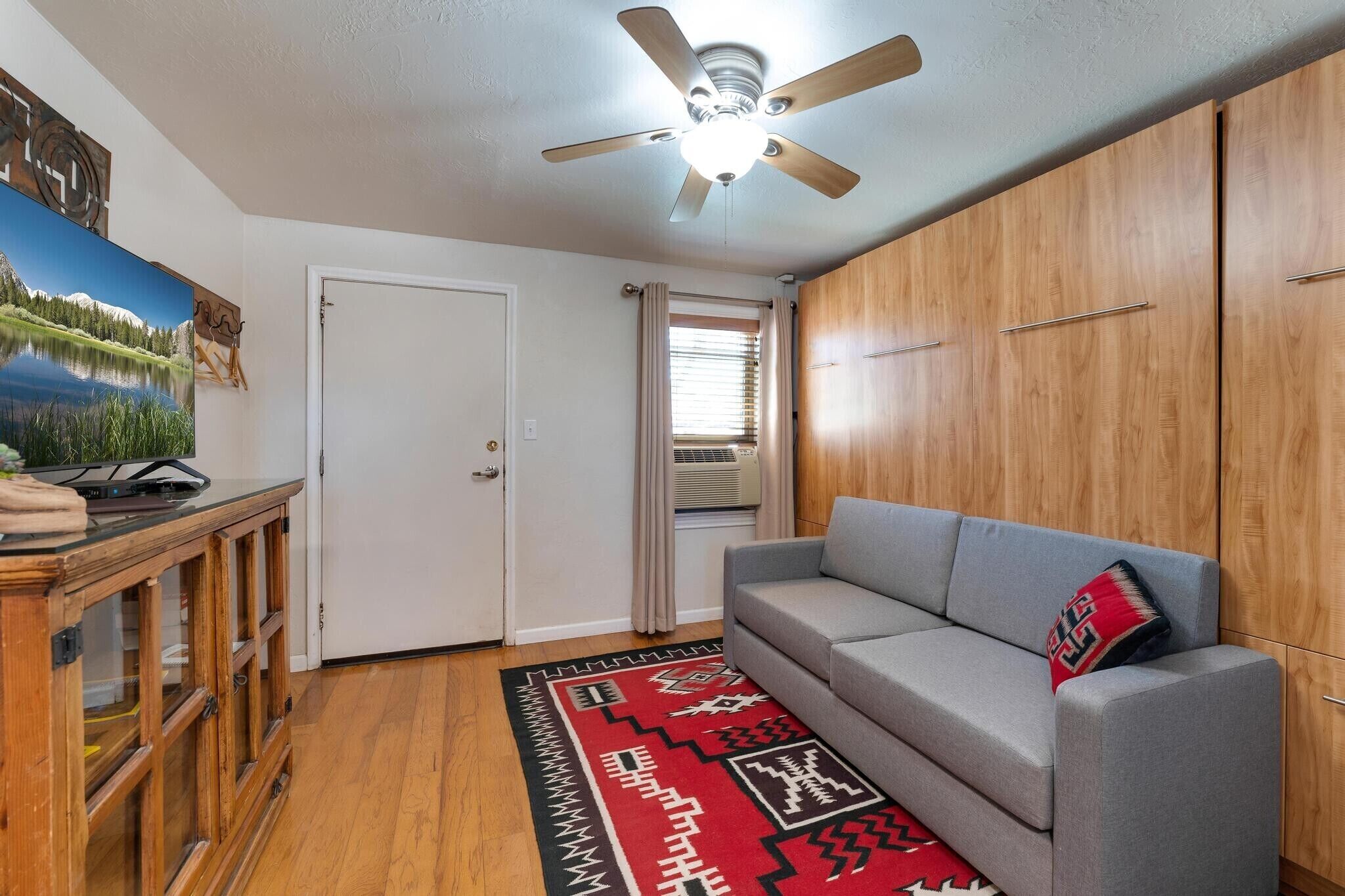 A room with a ceiling fan, a grey sofa with a red pillow, a wooden cabinet, a wooden desk with a computer, and a red and black patterned rug.