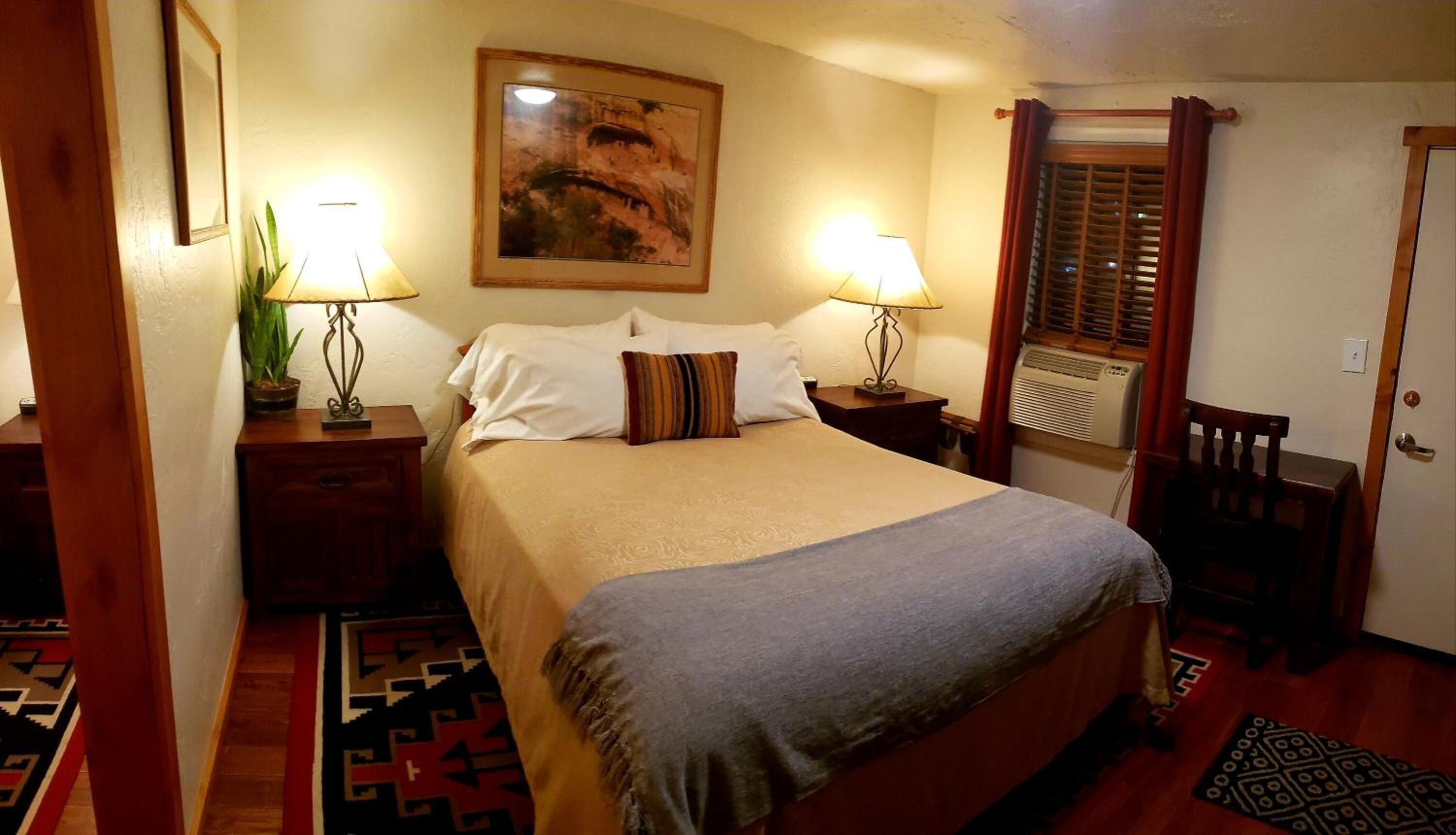 A neatly arranged bedroom with a bed, two bedside tables with lamps, a wall-mounted painting, a window with blinds, and a door.