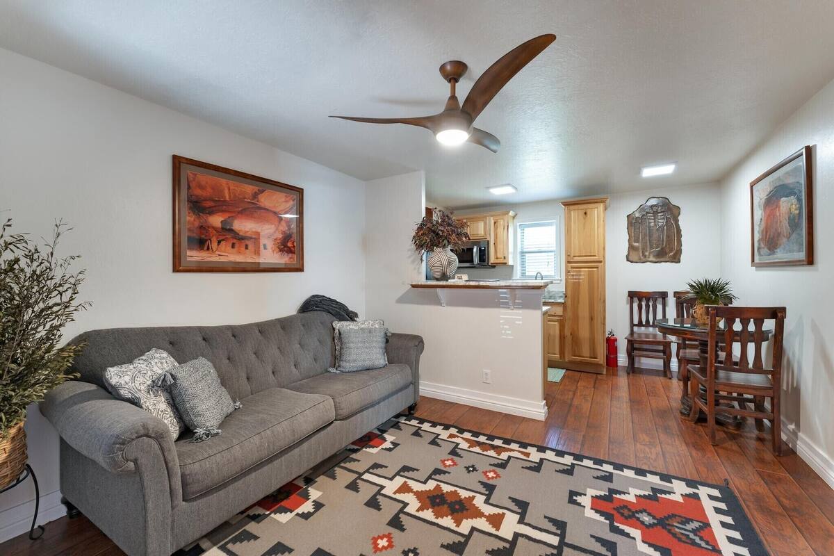 A living room with a gray sofa, a patterned rug, wooden dining chairs, a ceiling fan, and a kitchen in the background.