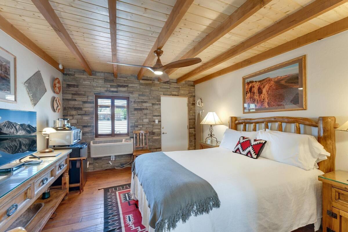 A bedroom with a stone wall, wooden ceiling beams, a bed with white linens, a desk with a lamp, and a painting on the wall.