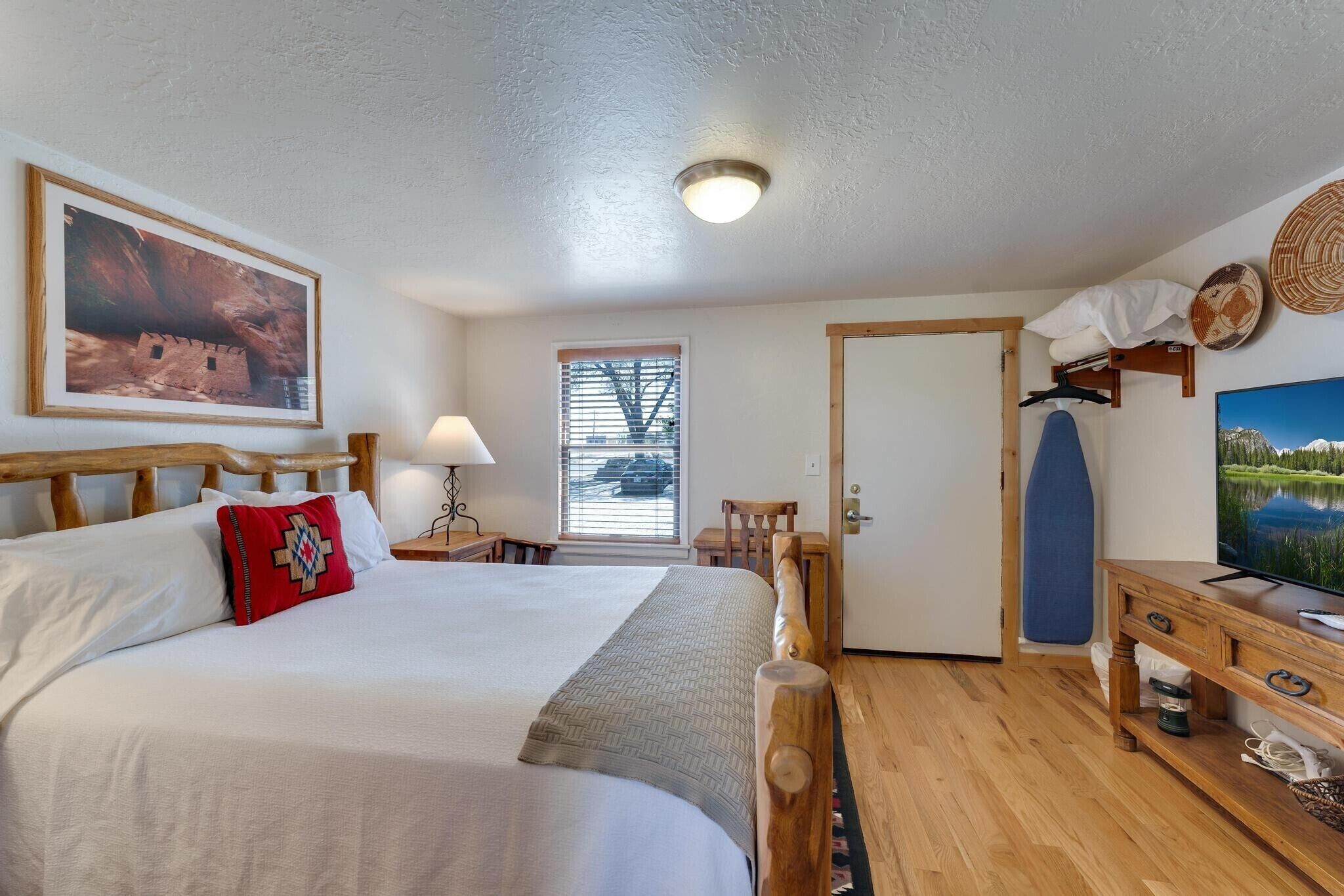A hotel room with a large bed, a television, a wooden headboard, a window with blinds, and a door.