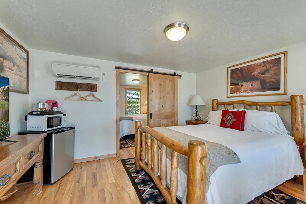 A room with a bed, a desk with a microwave, a small refrigerator, and a sliding door leading to another room.