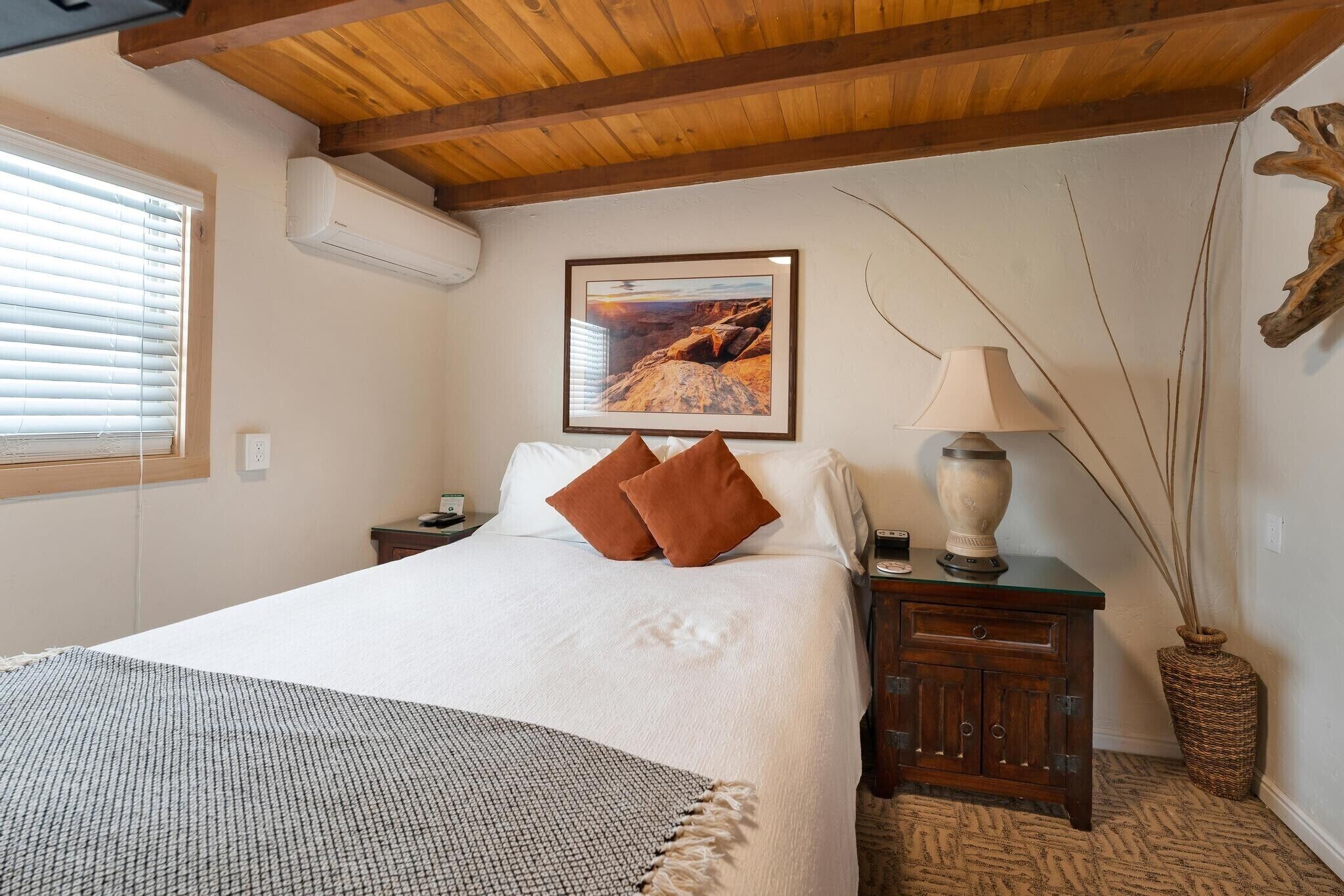 A bedroom with a large bed, a wooden ceiling, a framed picture on the wall, a bedside table with a lamp, and a vase with long stems.
