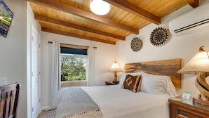 A bedroom with a wooden ceiling, a bed with white linens, two wall decor pieces, a nightstand with a lamp, and a window with curtains.