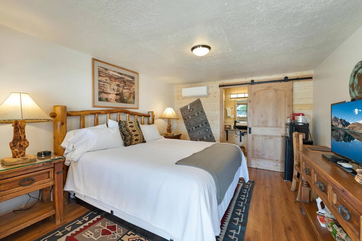 A bedroom with a large bed, wooden headboard, two bedside lamps, a framed picture on the wall, and a television on a wooden stand.