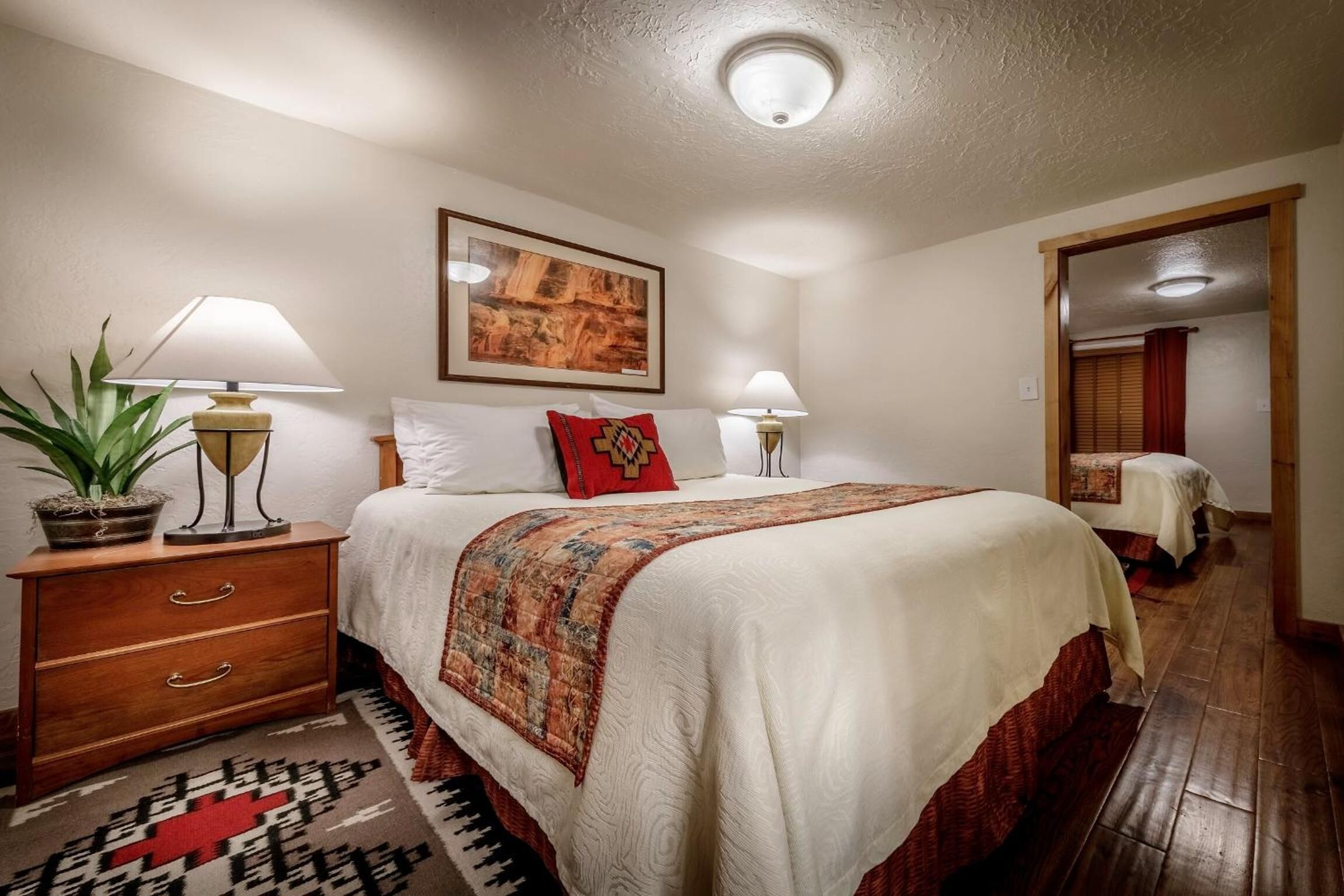 A well-lit bedroom with a large bed, two bedside tables with lamps, a potted plant, and a framed artwork on the wall.