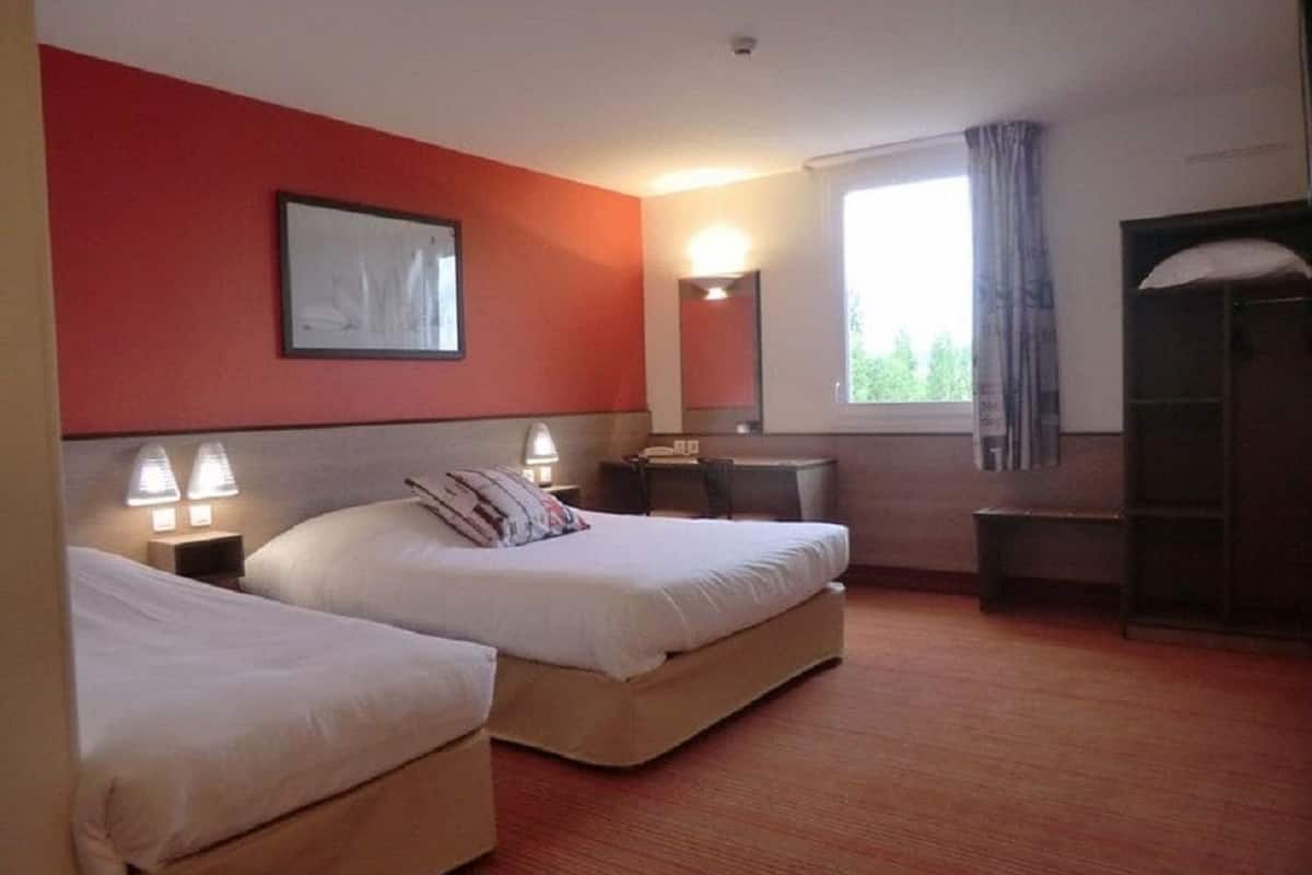 A hotel room with two beds, a desk, a chair, a window with curtains, and a mirror.