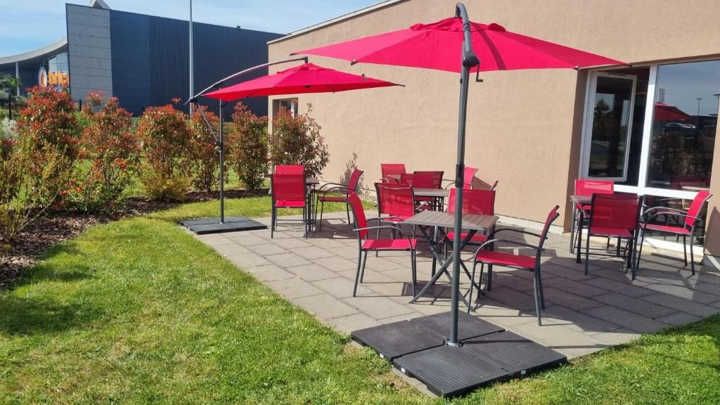 Un patio exterior con sillas rojas y sombrillas, un edificio con entrada de vidrio y un área verde con arbustos.