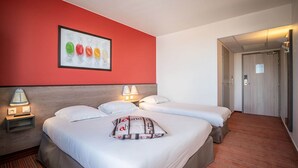 Une chambre d’hôtel avec deux lits, un mur d’accent rouge, une œuvre d’art encadrée et une porte donnant sur un couloir.
