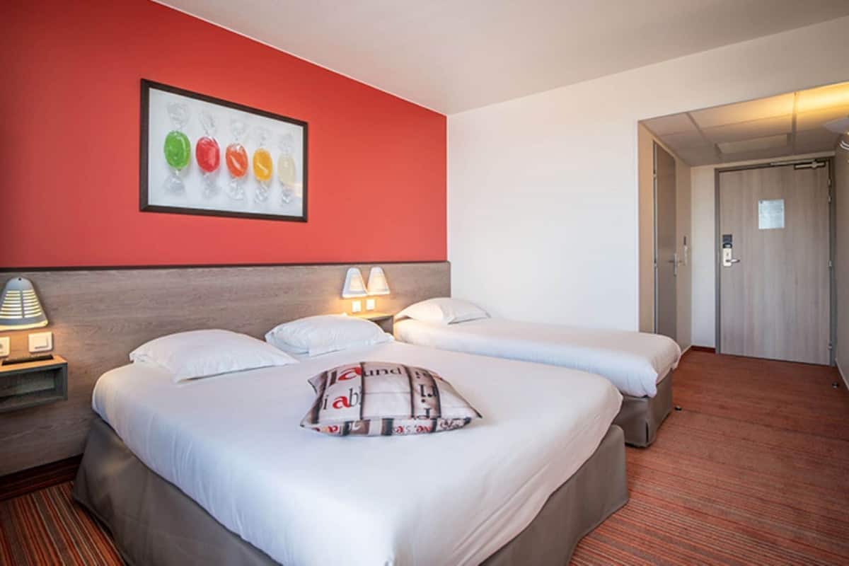 A hotel room with two beds, a red accent wall, a framed artwork, and a door leading to a hallway.