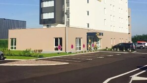 Un hôtel moderne, reconnaissable à son grand panneau « ACE » placé en haut à gauche, à sa façade sobre et à son parking situé devant l’entrée.