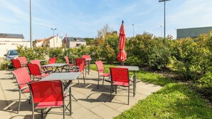 Une terrasse extérieure avec des chaises et des tables rouges, entourée de verdure et avec des bâtiments en arrière-plan.