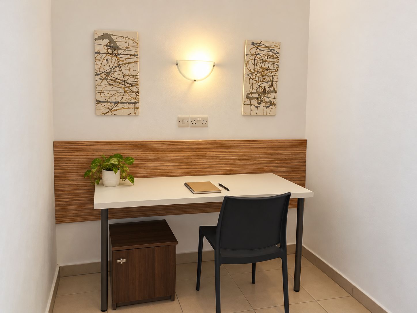 Un espacio de trabajo moderno con un escritorio, una silla y un pequeño armario. Hay dos cuadros abstractos en la pared sobre el escritorio.