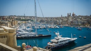 Un puerto deportivo con varias embarcaciones amarradas, un velero destacado y un paisaje urbano al fondo.
