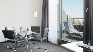 A modern hotel room with a glass table, four chairs, a sofa, a balcony with a chair, and a view of the city. - Living Hotel Frankfurt (Frankfurt)