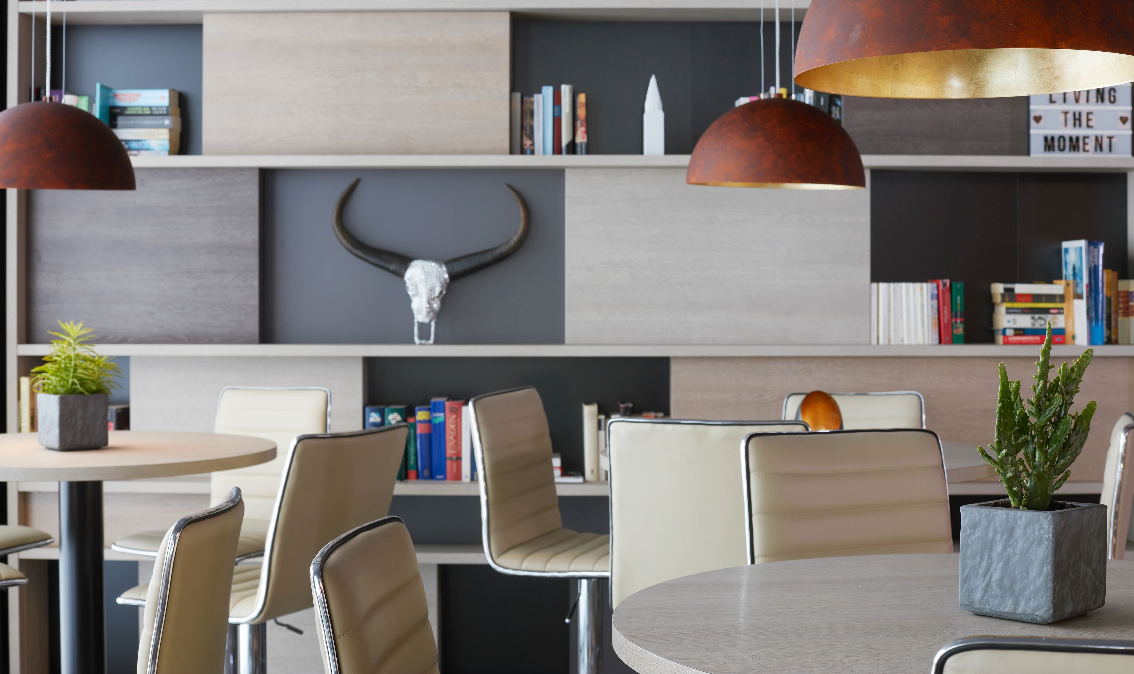 Ein modernes Interieur mit einem runden Tisch, beigen Stühlen, einer Topfpflanze und einem Bücherregal mit Büchern und einem dekorativen Schädel.