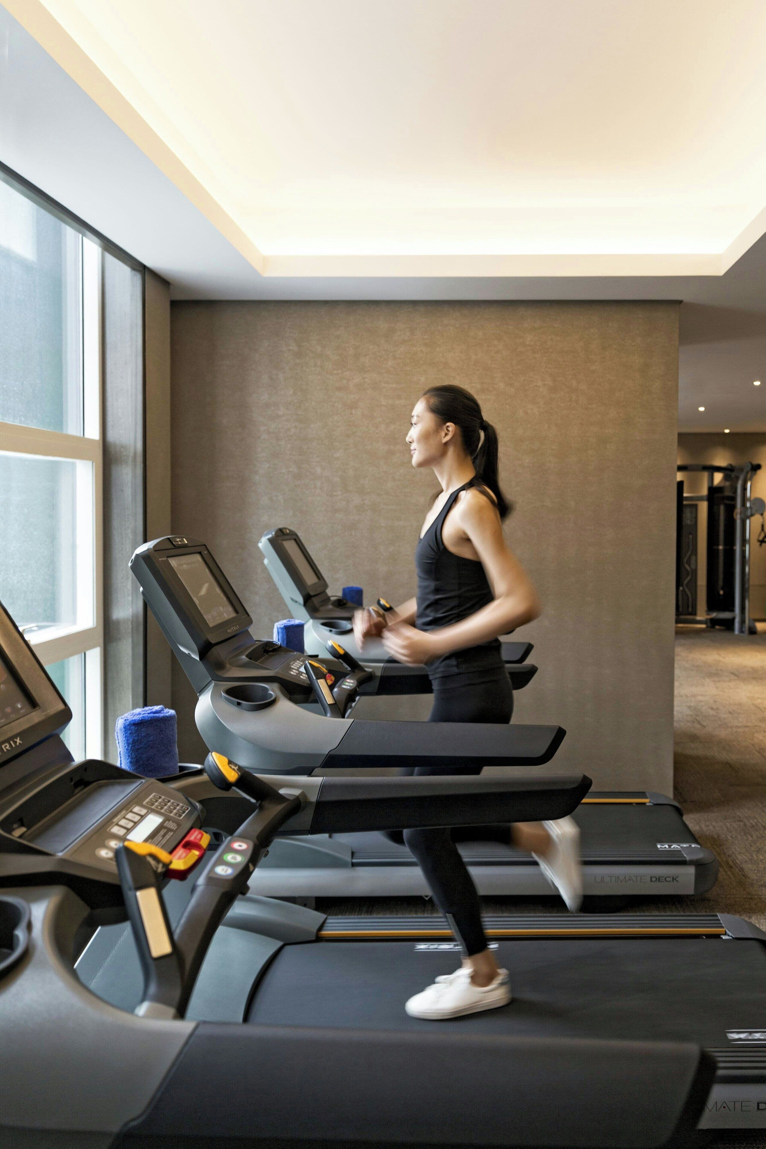 A woman running on a treadmill in a modern hotel gym with large windows and a neutral color palette.