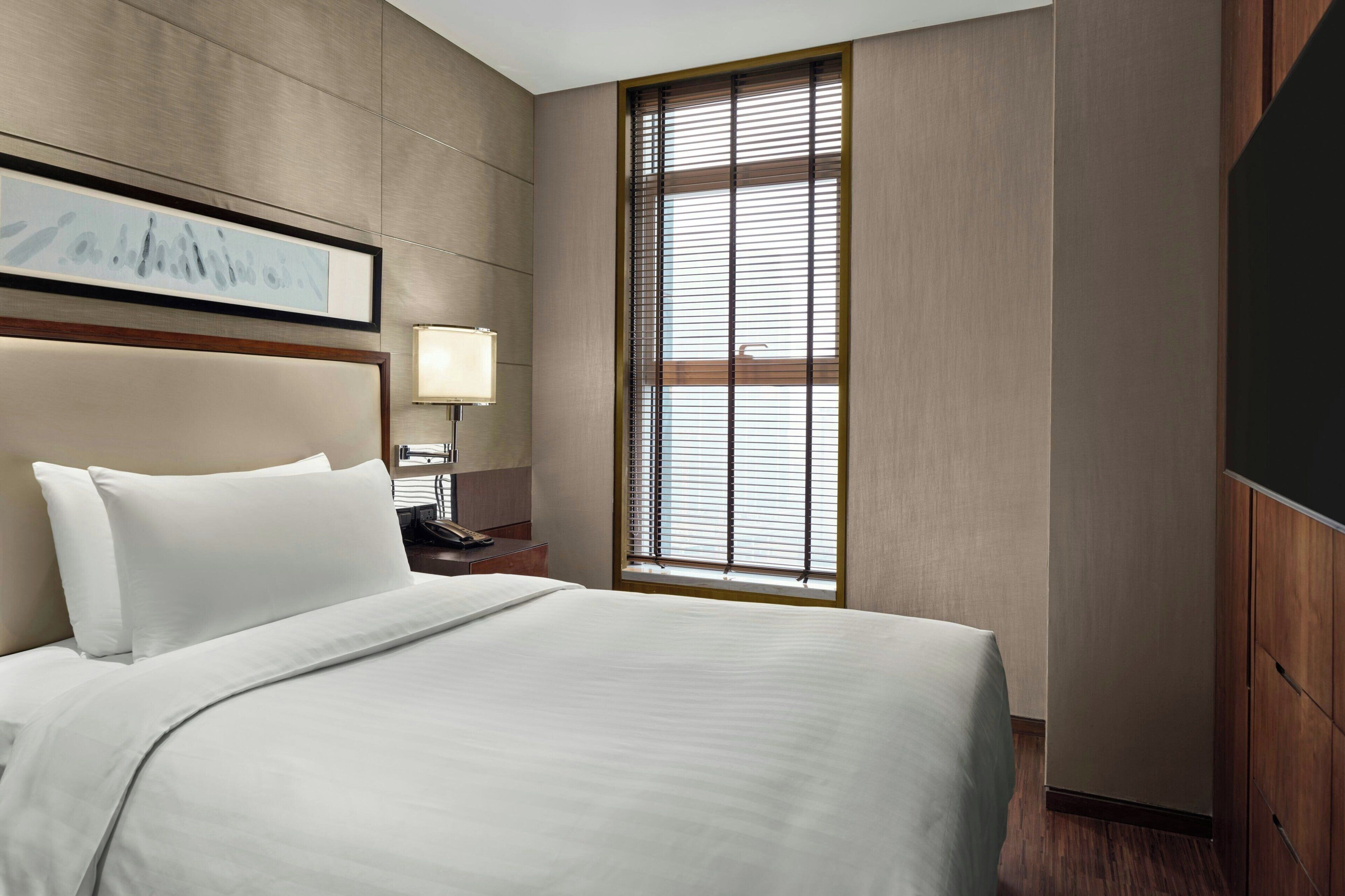 Une chambre d’hôtel avec un grand lit, une table de chevet avec une lampe, un tableau encadré au mur et une fenêtre avec des stores.