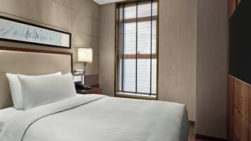 Une chambre d’hôtel avec un grand lit, une table de chevet avec une lampe, un tableau encadré au mur et une fenêtre avec des stores.