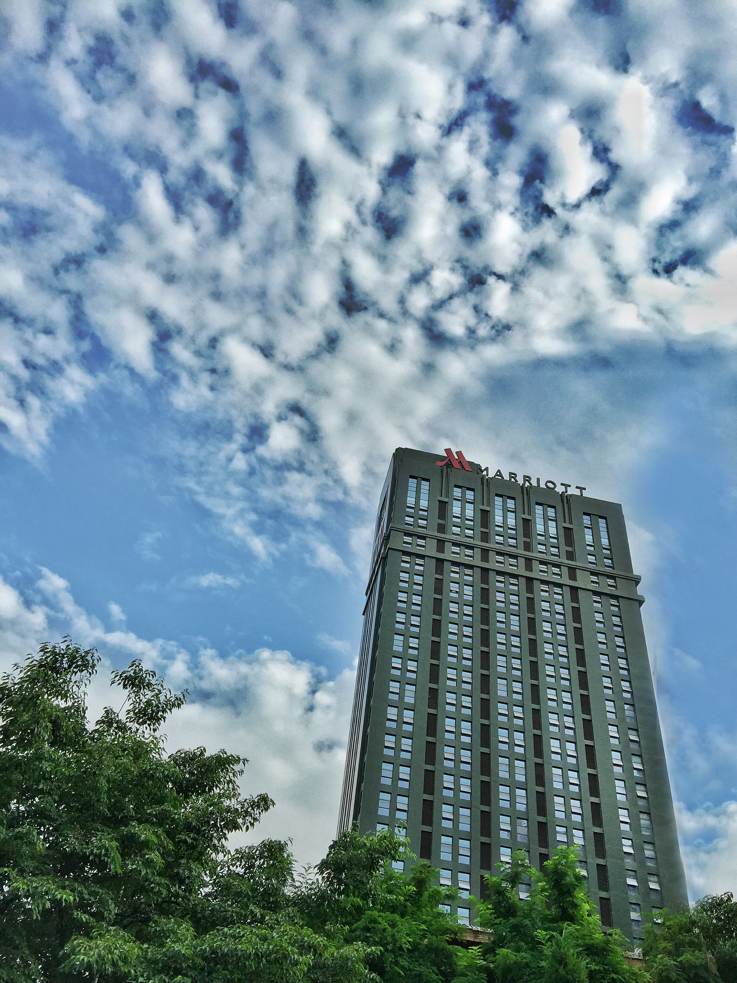 A tall hotel building with a visible 'H' logo on the top, surrounded by trees and a partly cloudy sky.