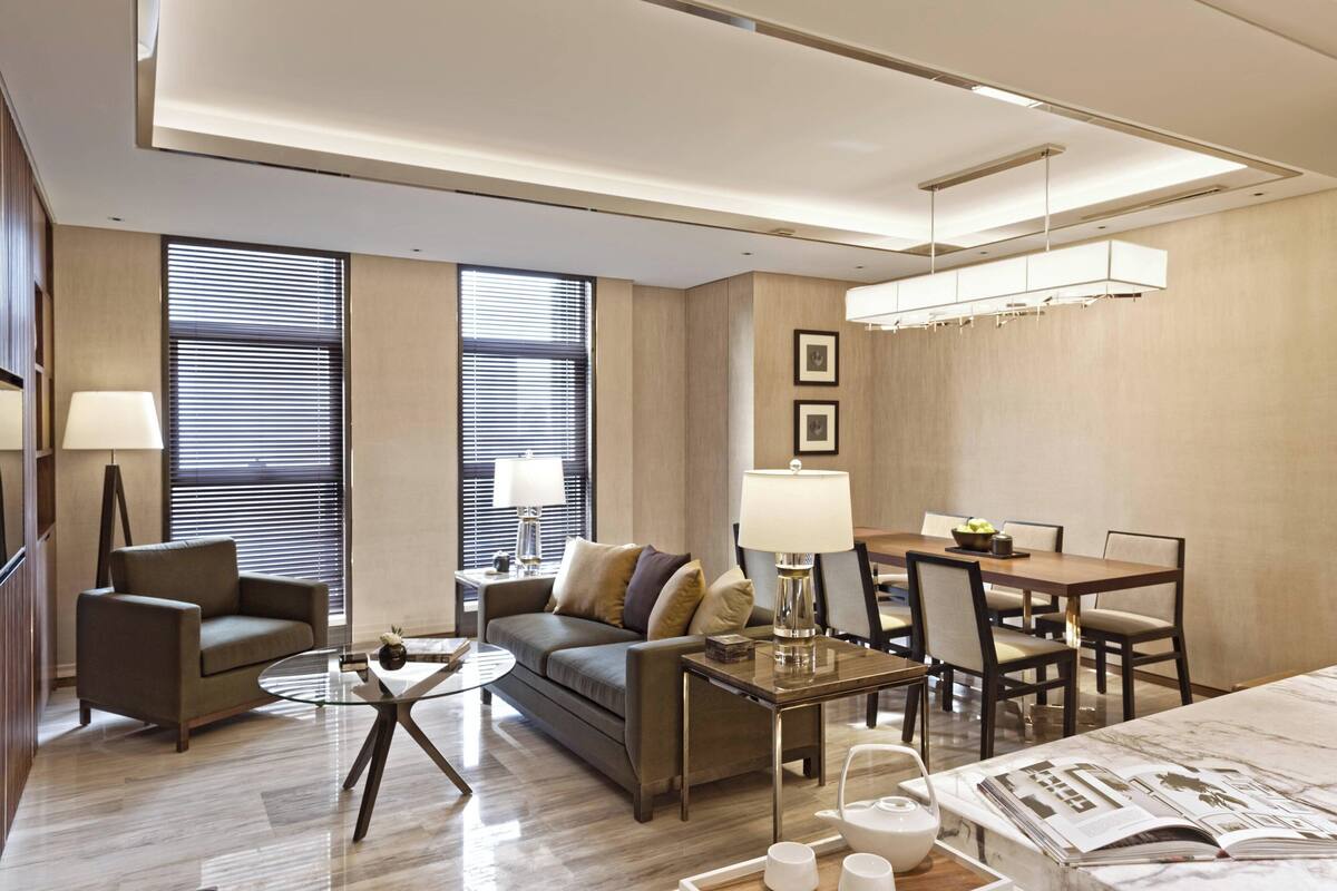 A modern hotel room with a sofa, armchairs, a dining table, and a coffee table. There are two windows with blinds, a lamp, and a magazine on the table.